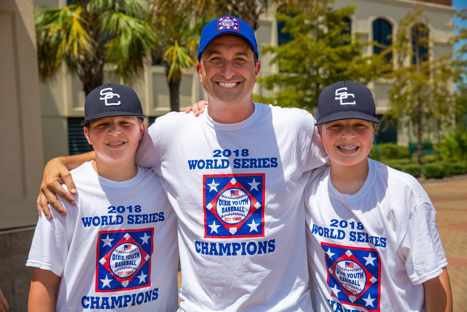 A man and two boys in 2018 Dixie Youth Baseball World Series champions shirts smile at a North Charleston celebration for the team.