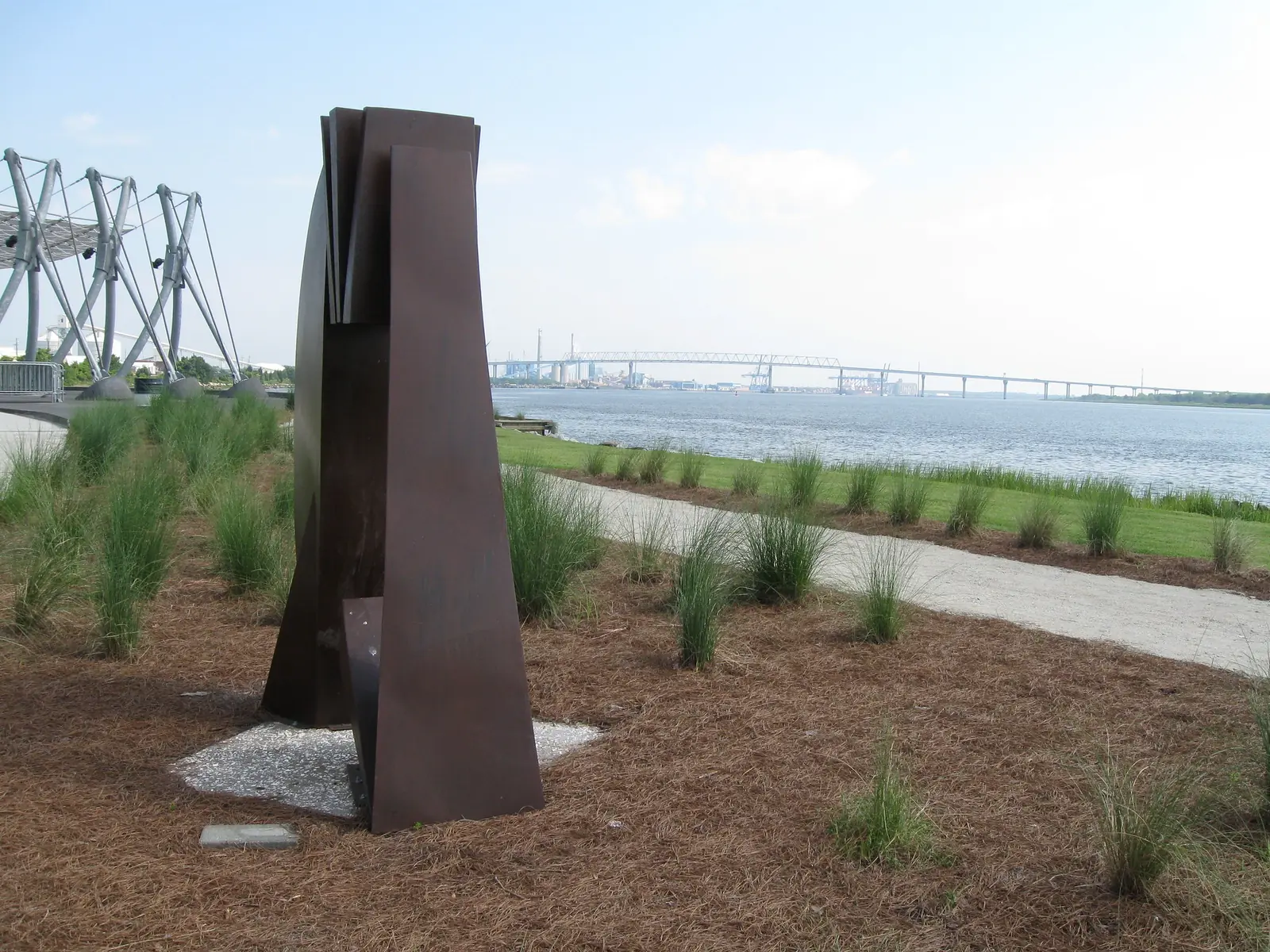 A tall steel sculpture stands beside a riverside path at Riverfront Park in North Charleston, with the Cooper River beyond.