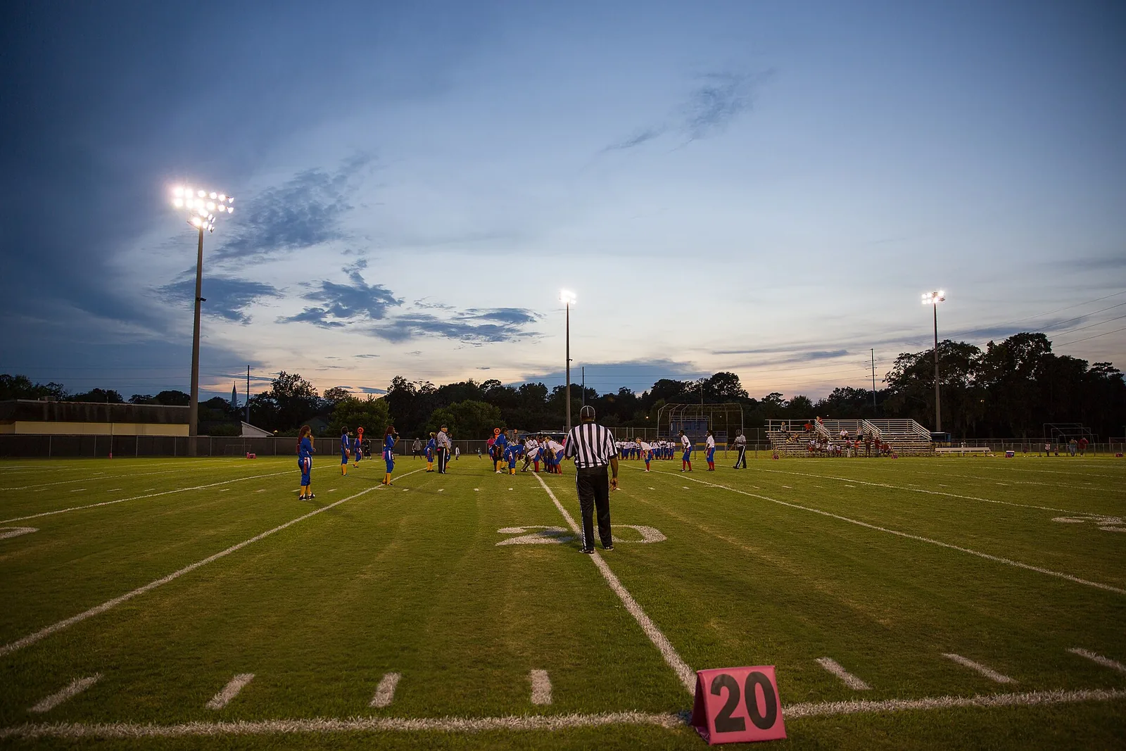 Stadium lights come on over a football field in North Charleston at dusk.