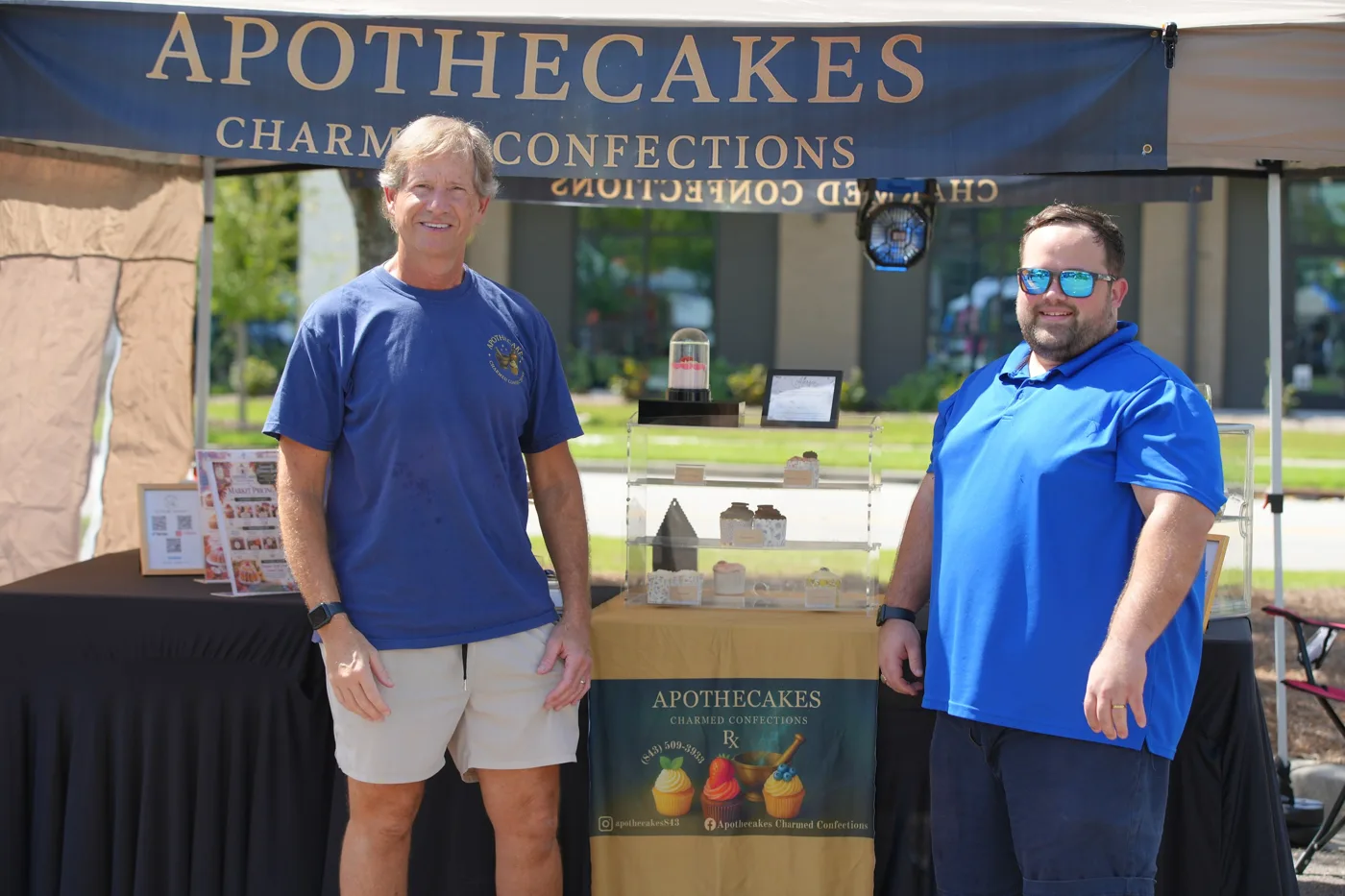 Two men stand in front of the Apothecakes Charmed Confections booth under a navy banner.