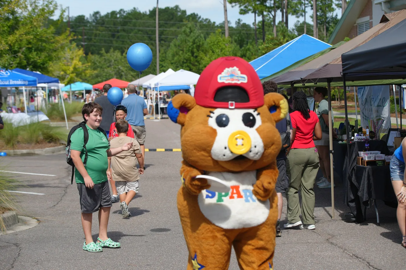 A costumed bear mascot in a red cap and a SPARK shirt walks between the vendor tents.