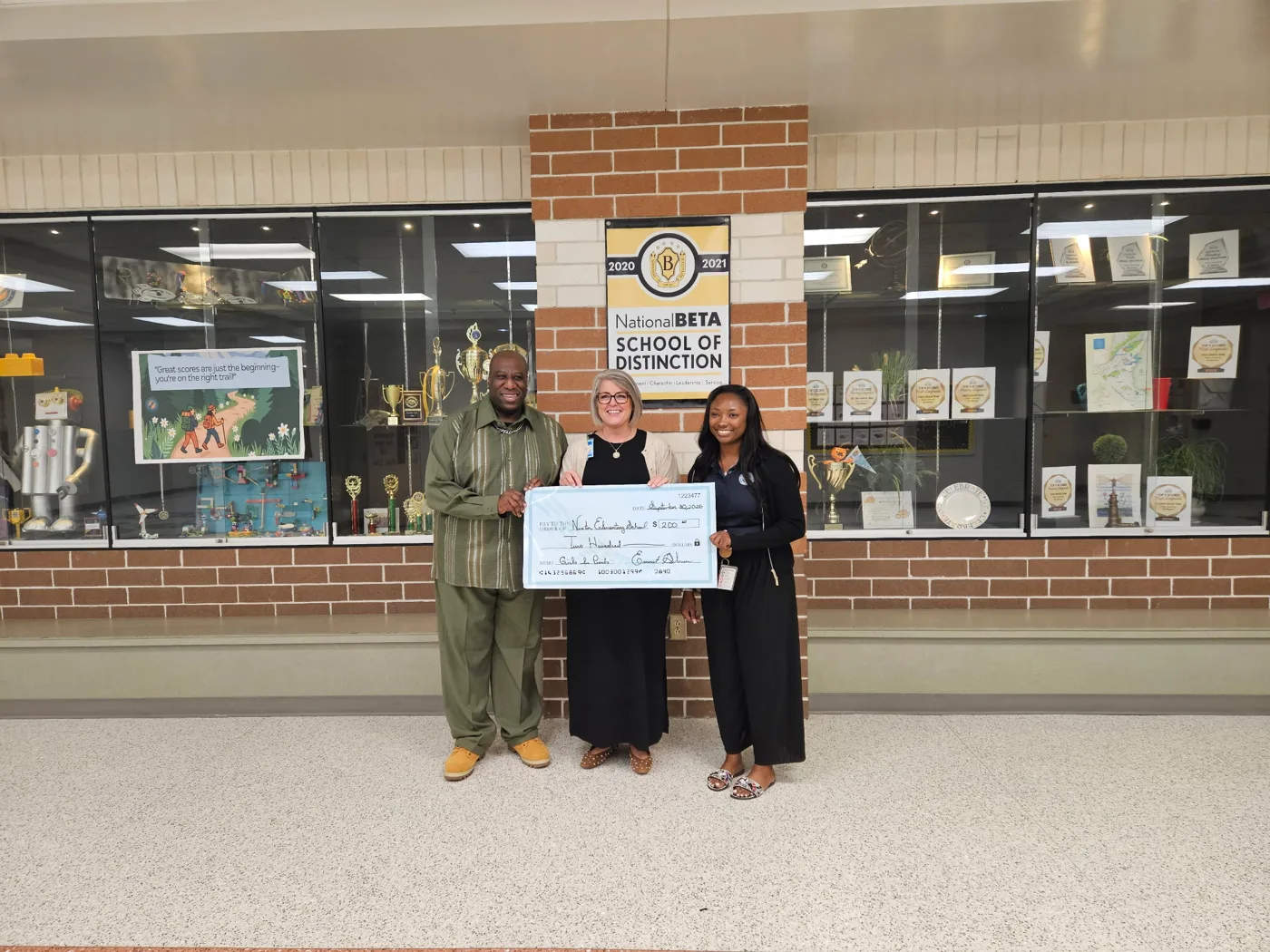 Three people hold an oversized check in the front hall of Nexton Elementary School.